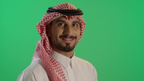 الوقوف بأيدى متقاطعة, رجل عربي خليجي سعودي يرتدي الثوب والشماغ ينظرإلى الكاميرا بايمائات السرور, خلفية خضراء، كروما