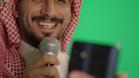 التواصل الفعال وتبادل الحوار و النقاش، بورتريه لشاب عربي خليجي سعودي يرتدي الثوب التقليدي والشماغ  يحمل بيده مكبر الصوت و الهاتف يقوم بإلقاء الخطاب بإيماءات الاندماج، إقامة ورشة عمل ودورة تدريبية، خلفية خضراء، كروما