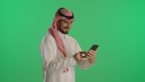 A portrait of a smiling Saudi Gulf Arab man wearing traditional attire and a shemagh, holding a mobile phone in his hand, chatting with family, using modern and advanced technology, with a green background, chroma.