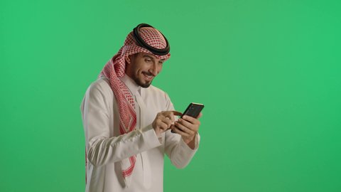 تصفح وسائل التواصل الاجتماعي عبر الجوال, استخدام الأجهزة التقنية الحديثة و المتطورة, بورتريه لرجل عربي خليجي سعودي مبتسم يرتدي الثوب التقليدي و الشماغ يمسك الهاتف المحمول بيده، خلفية خضراء، كروما