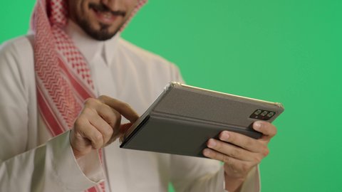 قضاء وقت ممتع، تصفح الإنترنت وإنجاز العمل عن بعد، بورتريه لرجل عربي خليجي سعودى يرتدى الثوب التقليدى و الشماغ يجلس على كرسي مريح يستخدم جهاز التابلت ، ايماءات السعاده و السرور، خلفية خضراء، كروما