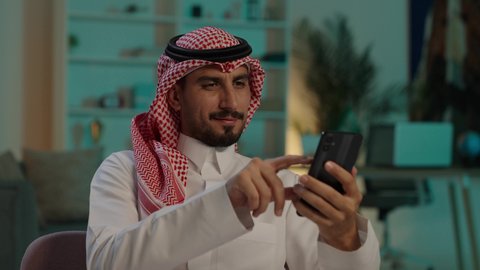 التسوق الالكترونى عبر الانترنت، رجل عربي خليجي سعودي يرتدي الثوب التقليدي والشماغ  يمسك الهاتف بيده يجلس على الاريكة، تصفح وسائل التواصل الاجتماعي عبر الجوال