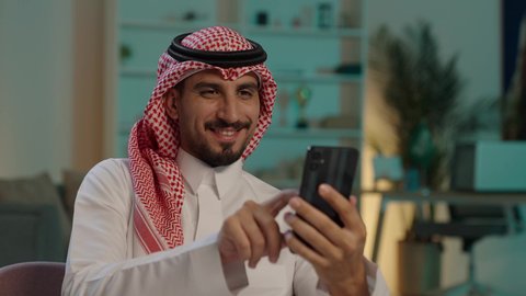 مفهوم الجلوس والراحة، رجل عربي خليجي سعودي يرتدي الثوب التقليدي والشماغ  يمسك الهاتف المحمول بيده يجلس على الاريكة، تصفح وسائل التواصل الاجتماعي عبر الهاتف