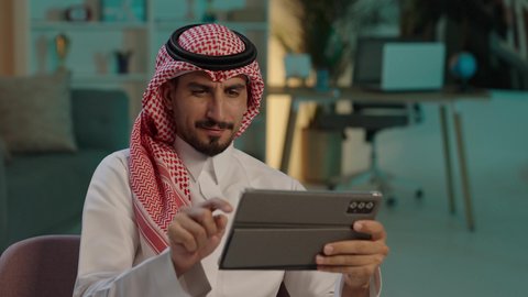 التسوق الإلكتروني الآمن، قضاء الوقت بتصفح برامج التواصل الاجتماعي، رجل عربي خليجي سعودى يرتدى الثوب التقليدى و الشماغ يجلس على كرسي مريح يستخدم جهاز التابلت ، ايماءات السعاده و السرور