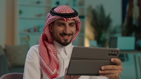 قضاء الوقت بتصفح برامج التواصل الاجتماعي، رجل عربي خليجي سعودى يرتدى الثوب التقليدى و الشماغ يجلس على كرسي مريح يستخدم جهاز التابلت ، التسوق الإلكتروني الآمن، ايماءات السعاده و السرور