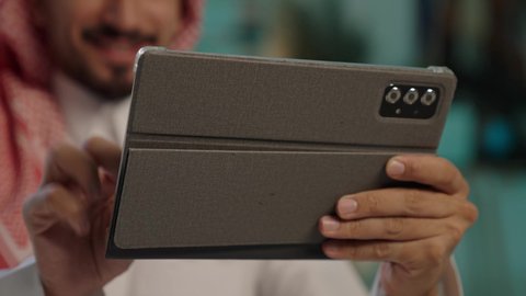 دمج التكنولوجيا في الحياة العملية، تصفح الإنترنت وإنجاز العمل عن بعد، بورتريه لرجل عربي خليجي سعودى يرتدى الثوب التقليدى و الشماغ يجلس على كرسي مريح يستخدم جهاز التابلت ، ايماءات السعاده و السرور، خلفية خضراء، كروما