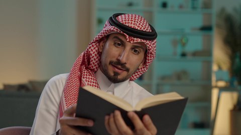 تنمية المهارات الفكرية ، الاستمتاع بالهدوء والراحة داخل المنزل، قضاء الوقت بالمطالعة والثقافة، ممارسة الهوايات المفضلة، رجل عربي خليجي سعودي يرتدي الزى التقليدي و الشماغ يجلس على الأريكة يقوم بقراءة كتاب