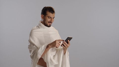 استخدام خدمة تطبيق الارشادات لأداء مناسك الحج والعمرة، بورتريه لرجل مسلم خليجي عربي سعودي مبتسم يرتدي ملابس الإحرام يستخدم الهاتف المحمول، تطبيق مرشد الحج الالكتروني، مفهوم أركان الإسلام، خلفية رمادية
