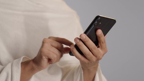 بورتريه مقرب ليد لرجل عربي خليجي سعودي يرتدي ملابس الاحرام يستخدم الهاتف المحمول، زيارة بيت الله الحرام في مكة المكرمة، الركن الخامس من أركان الاسلام، آداء مناسك الحج والعمرة، خلفية رمادية