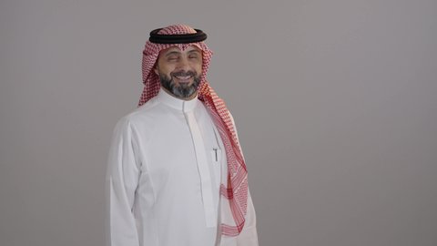 الشعور بالثقة في النفس، الاهتمام بالأناقة والشياكة والمظهر الخارجي، بورتريه لرجل عربي خليجي سعودى مبتسم يرتدى الثوب التقليدى والشماغ ينظر إلى الكاميرا يقف بأيدي متقاطعة، مفهوم الرجولة والاناقة، خلفية رمادية