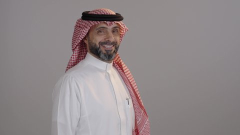 بورتريه لرجل عربي خليجي سعودى مبتسم يرتدى الثوب التقليدى والشماغ ينظر إلى الكاميرا يقف بأيدي متقاطعة، مفهوم الرجولة والاناقة، الشعور بالثقة في النفس، الاهتمام بالأناقة والشياكة والمظهر الخارجي، خلفية رمادية