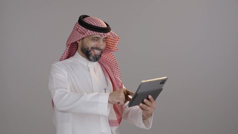ايماءات السعادة والسرور، الدردشة والتحدث مع الاصدقاء عبر الانترنت، بورتريه لرجل عربي خليجي سعودى يرتدى الثوب التقليدى والشماغ  يقوم بمتابعة اخبار وسائل التواصل الاجتماعي باستخدام التابلت، تصفح تطبيقات جهاز التابلت، استخدام الأجهزة التقنية الحديثة، خلفية رمادية