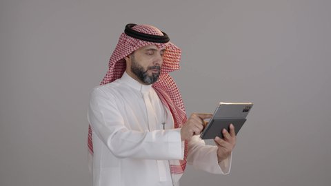 بورتريه لرجل عربي خليجي سعودى يرتدى الثوب التقليدى والشماغ  يقوم بمتابعة اخبار وسائل التواصل الاجتماعي باستخدام التابلت، ايماءات السعادة والسرور، الدردشة والتحدث مع الاصدقاء عبر الانترنت، تصفح تطبيقات جهاز التابلت، استخدام الأجهزة التقنية الحديثة، خلفية رمادية