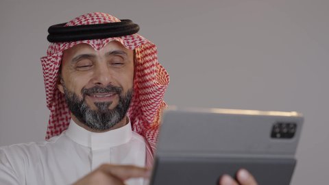 النظر الى الكاميرا بايماءات السعادة والسرور، بورتريه لرجل عربي خليجي سعودى يرتدى الثوب التقليدى والشماغ يقوم بمتابعة اخبار وسائل التواصل الاجتماعي باستخدام التابلت، تصفح تطبيقات جهاز التابلت، استخدام الأجهزة التقنية الحديثة، خلفية رمادية