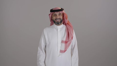 The feeling of satisfaction and approval, expressing admiration and appreciation, raising the hand with signs of quality and acceptance, a portrait of a smiling Saudi Arabian Gulf man wearing traditional attire and a shemagh, raising his hand upwards, looking at the camera with gestures of happiness and joy, the concept of masculinity and elegance, gray background.