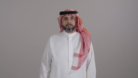 Expressions of rejection, disapproval, and dissatisfaction, anxiety and failure to achieve something, the concept of negative evaluation and discomfort, close-up shot, a portrait of a Saudi Arabian Gulf man wearing traditional attire and a ghutrah looking at the camera, pointing his thumb down with gestures of discomfort, gray background.