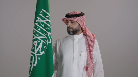 مفهوم الولاء وحب الوطن والفخربالانتماء للسعودية، بورتريه لرجل عربي خليجي سعودى مبتسم يرتدى الثوب التقليدى والشماغ يمسك علم المملكة بإيماءات الاعتزاز، إحياء ذكرى اليوم الوطني السعودي، المشاركة في الاحتفال بالمناسبات والأعياد الوطنية، خلفية رمادية
