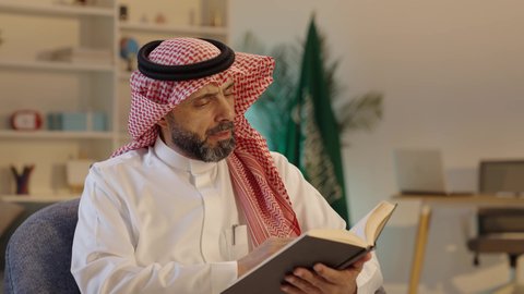 Spending free time on education and learning, reading and studying is a daily habit, the culture of self-learning, the interest in browsing useful books, an Arab Gulf Saudi man wearing traditional attire and a shemagh sitting on a sofa at home holding a book in his hand.