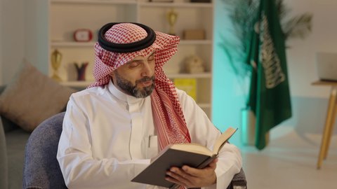 Reading and studying is a daily habit, a culture of self-learning, developing reading skills, spending free time on education and learning, and being interested in browsing useful books. An Arab Gulf Saudi man wearing traditional attire and a headscarf is sitting on a chair at home holding a book in his hand.