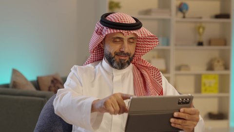 الدردشة والتحدث مع الاصدقاء عبر الانترنت، رجل عربي خليجي سعودى يرتدى الثوب التقليدى والشماغ  يقوم بمتابعة اخبار وسائل التواصل الاجتماعي باستخدام التابلت، تصفح تطبيقات جهاز التابلت، استخدام الأجهزة التقنية الحديثة، ايماءات السعادة والسرور