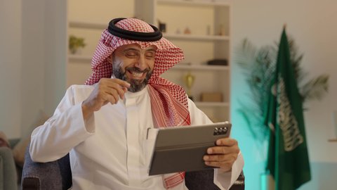 تصفح تطبيقات جهاز التابلت، الدردشة والتحدث مع الاصدقاء عبر الانترنت، استخدام الأجهزة التقنية الحديثة، رجل عربي خليجي سعودى يرتدى الثوب التقليدى والشماغ  يقوم بمتابعة اخبار وسائل التواصل الاجتماعي باستخدام التابلت، ايماءات السعادة والسرور