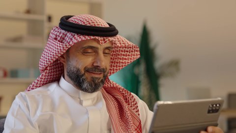 تصفح مواقع الانترنت، ايماءات السعادة والسرور،استخدام الأجهزة التقنية الحديثة، رجل عربي خليجي سعودى يرتدى الثوب التقليدى والشماغ يستخدم التابلت، الدردشة مع الأهل والأصحاب