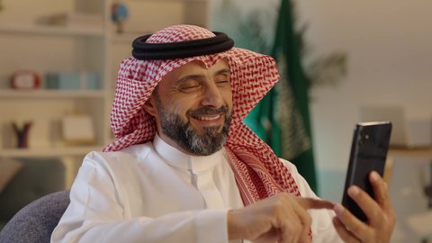 التواصل مع الأصدقاء، رجل عربي خليجي سعودى مبتسم يرتدى الثوب التقليدى والشماغ  يجلس على الكرسى يمسك الهاتف المحمول، استخدام أجهزة تقنية حديثة