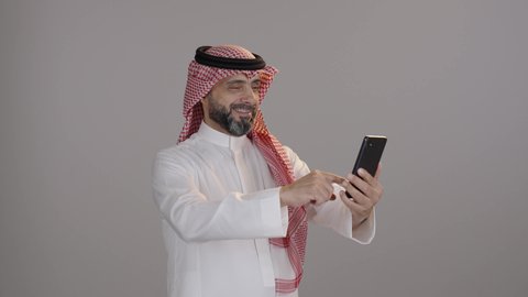 Online shopping using a phone, spending time browsing social media, completing tasks remotely, using modern technological devices, a portrait of an Arab Saudi Gulf man wearing a thobe and shemagh using a mobile phone, gray background.