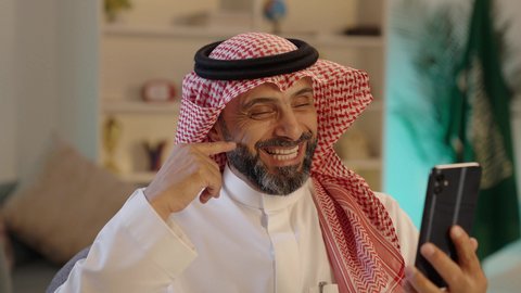 مكالمات فيديو للتواصل الاجتماعي عبر الجوال، ايماءات تدل على السعادة، استخدام التكنولوجيا و التقنيات الحديثة، رجل عربي خليجي سعودى مبتسم يرتدى الثوب التقليدى والشماغ  يجلس على الكرسى يمسك الهاتف المحمول