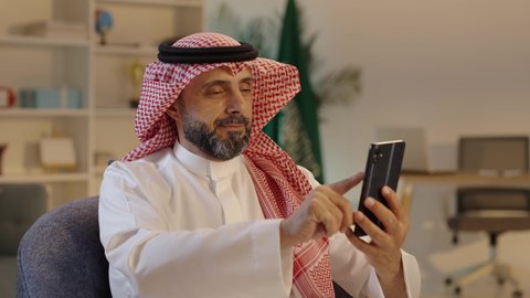 التسوق الالكتروني من خلال الجوال، رجل عربي خليجي سعودي    يرتدي الثوب والشماغ يجلس على كرسى يستخدم الجوال، استعمال أجهزة تقنية حديثة