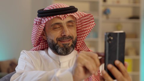 رجل عربي خليجي سعودى مبتسم يرتدى الثوب التقليدى والشماغ  يجلس على الكرسى يمسك الهاتف المحمول ،التواصل مع الأصدقاء ،استخدام أجهزة تقنية حديثة