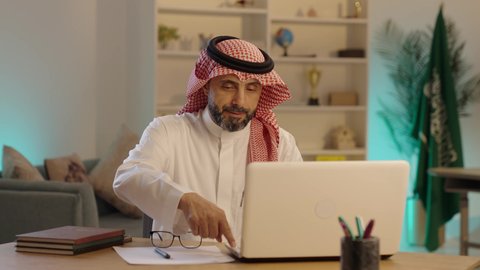 تدوين الملاحظات على الورق، إدارة شؤون الأعمال وتنظيمها، دمج العمل مع التقنية والتكنولوجيا، تطور الشركات السعودية، استخدام جهاز تقني حديث ومتطور، رجل عربي خليجي سعودى يرتدى الثوب التقليدى والشماغ و انظارة الطبية يستخدم الحاسوب، مهنة ووظيفة مكتبية