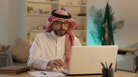 انجاز مهام الأعمال عن بعد، رجل عربي خليجي سعودى يرتدي الثوب التقليدى والشماغ  والنظارة يعمل على جهاز الحاسوب المحمول و يكتب شيء ما، دمج التقنيات الحديثة والمتطورة في الحياة العملية، مفهوم ادارة الاعمال عن بعد، مهن ووظائف مكتبيه سعودية