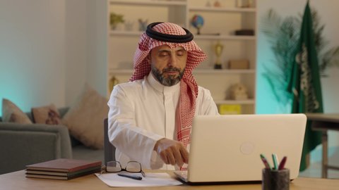إدارة شؤون الأعمال وتنظيمها، دمج العمل مع التقنية والتكنولوجيا، تطور الشركات السعودية، استخدام جهاز تقني حديث ومتطور، رجل عربي خليجي سعودى يرتدى الثوب التقليدى والشماغ و النظارة يستخدم الحاسوب، مهنة ووظيفة مكتبية
