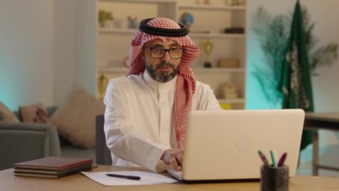 رجل عربي خليجي سعودى يرتدي الثوب التقليدى والشماغ  والنظارة يعمل على جهاز الحاسوب المحمول و يكتب شيء ما، دمج التقنيات الحديثة والمتطورة في الحياة العملية، مفهوم ادارة الاعمال عن بعد، مهن ووظائف مكتبيه سعودية