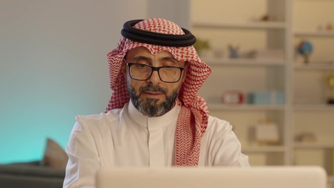 استخدام جهاز الحاسوب المحمول لإنجاز مهام الاعمال باحترافية، رجل عربي خليجي سعودي يرتدي الثوب والشماغ التقليدي و النظارة يجلس على المكتب يقوم بالعمل عبر اللابتوب، ادارة الأعمال عن بعد