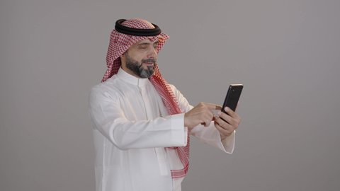 بورتريه لرجل عربي خليجي سعودي يرتدي الثوب والشماغ يستخدم الجوال، التسوق عبرالانترنت باستخدام الهاتف, قضاء الوقت بتصفح برامج التواصل الاجتماعي, إنجاز المهام عن بعد، استعمال أجهزة تقنية حديثة، خلفية رمادية
