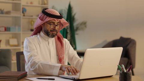 Reviewing plans, strategies, and agreements through modern technologies, using a laptop to accomplish business tasks professionally, an Arab Gulf Saudi man wearing traditional attire, a ghutrah, and glasses sitting at a desk working on a laptop, administrative and office jobs.