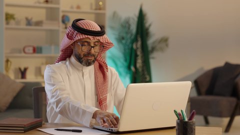استخدام جهاز الحاسوب المحمول لإنجاز مهام الاعمال باحترافية، رجل عربي خليجي سعودى يرتدي الثوب التقليدى والشماغ  والنظارة يجلس على مكتب يعمل على اللابتوب، مهن ووظائف ادارية مكتبية