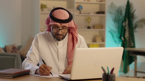 The use of modern devices and advanced technologies, the concept of remote business management, professions and office jobs in Saudi Arabia, an Arab Gulf Saudi man wearing traditional attire and a headscarf, sitting behind a desk taking notes next to a laptop.