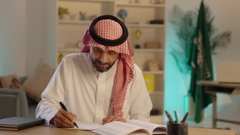 كتابة وتدوين الملاحظات المهمة، مفهوم ادارة الاعمال، مهن ووظائف مكتبيه في الممكلة العربية، رجل عربي خليجي سعودي يرتدي الثوب والشماغ التقليدي يجلس خلف طاولة المكتب بإيماءات الاندماج والتركيز