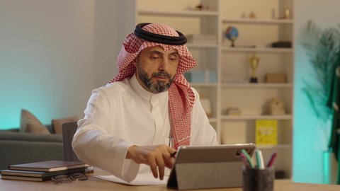 مفهوم ادارة الاعمال عن بعد، مهن ووظائف مكتبيه سعودية, رجل عربي خليجي سعودى يرتدي الثوب التقليدى والشماغ يجلس خلف طاولة خشبية يقوم بتدوين الملاحظات بجانب التابلت، استخدام الأجهزة الحديثة والتقنيات المتطورة