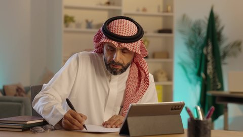 مهن ووظائف مكتبيه سعودية, مفهوم ادارة الاعمال عن بعد، رجل عربي خليجي سعودى يرتدي الثوب التقليدى والشماغ يجلس خلف طاولة خشبية يقوم بتدوين الملاحظات بجانب التابلت، استخدام الأجهزة الحديثة والتقنيات المتطورة