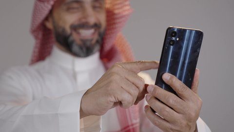 إستخدام أجهزة تقنية حديثة ، بورتريه مقرب لرجل سعودي عربي خليجي يرتدي الزي التقليدي السعودي يحمل بيده الهاتف المحمول ، التواصل والدردشة مع الأصدقاء ، تصفح الإنترنت ، ايماءات تدل على السعادة والسرور، خلفية رمادية