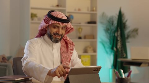قضاء الوقت بتصفح برامج التواصل الاجتماعي، رجل عربي خليجي سعودى كبير يجلس على كرسي مريح يستخدم جهاز التابلت ، التسوق الإلكتروني الآمن، استخدام اجهزة تقنية، ايماءات السعاده و السرور