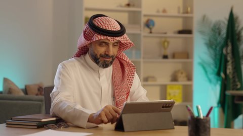 تصفح الإنترنت وإنجاز المهام عن بعد، رجل عربي خليجي سعودى كبير يجلس على كرسي مريح يستخدم جهاز التابلت ، التسوق الإلكتروني الآمن، الشراء والبيع عبر الاونلاين، النظر الى الكاميرا