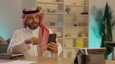 Making video calls via mobile phone, communicating with family and friends, using modern technology and techniques, an Arab Gulf Saudi man wearing traditional attire and a ghutrah using a mobile phone, gestures of happiness and joy.