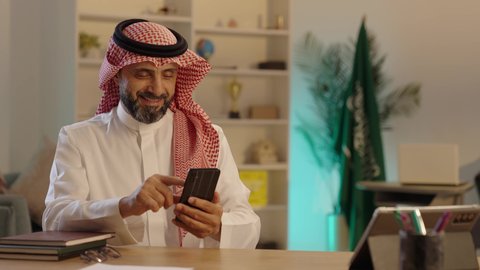Sitting in the living room, browsing the internet and following social media news through modern technologies, digital marketing online, communicating and chatting with friends via mobile phone, an Arab Gulf Saudi man wearing traditional attire and a shemagh is using his mobile phone.
