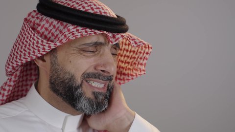 إيماءات الشعور بألم الأسنان، بورتريه لرجل عربي خليجي سعودى يرتدي الثوب التقليدي والشماغ يضع يديه على خده ويغمض عينيه, تسوس الأضراس وإلتهاب اللثه، طلب العلاج والمساعده، خلفية رمادية