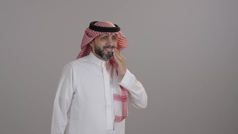 بورتريه لرجل عربي خليجي سعودى يرتدي الثوب التقليدي والشماغ يضع يديه على خده ويغمض عينيه بإيماءات الشعور بألم الأسنان, تسوس الأضراس وإلتهاب اللثه، طلب العلاج والمساعده، خلفية رمادية